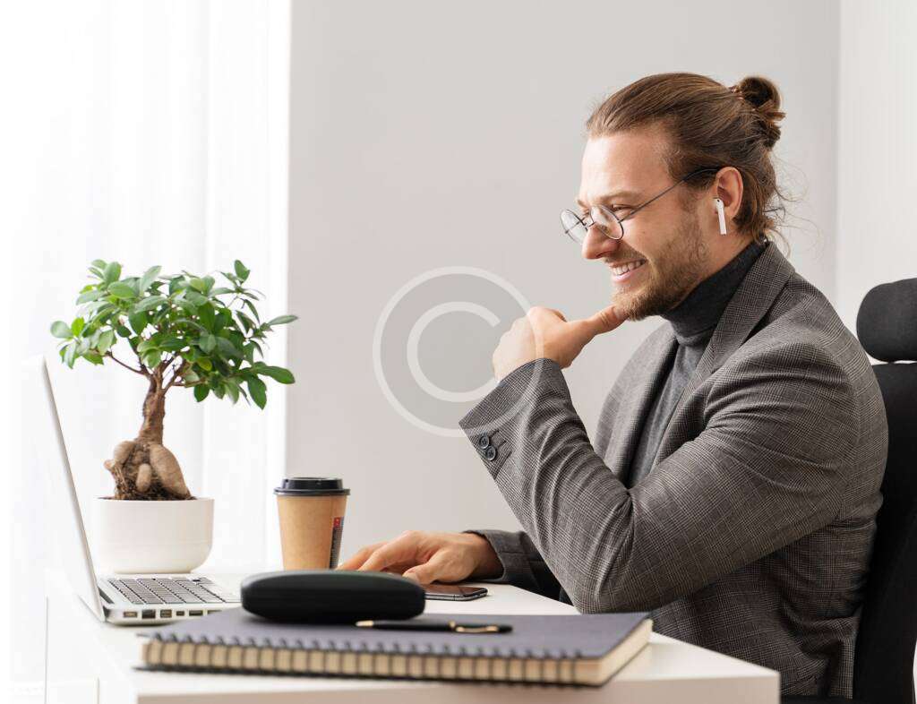 A man with a beard and glasses, wearing earbuds and a grey blazer, smiling while working on a laptop at a white desk with a bonsai tree and a coffee cup.