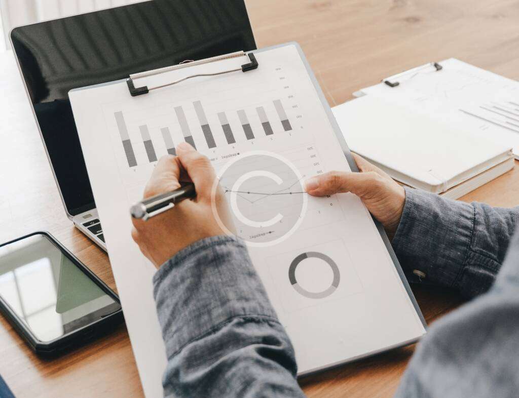 A person reviewing charts and graphs on a clipboard at a desk, with a laptop, smartphone, and documents in the background.