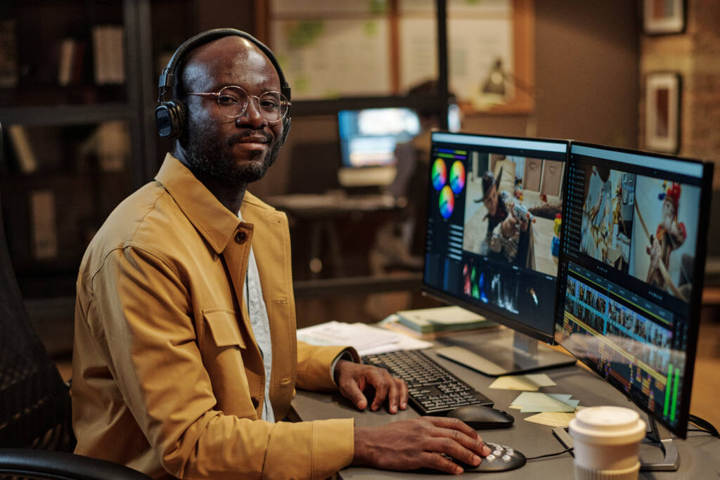 A video editor with headphones working on a video editing project on dual monitors in a dimly lit office.