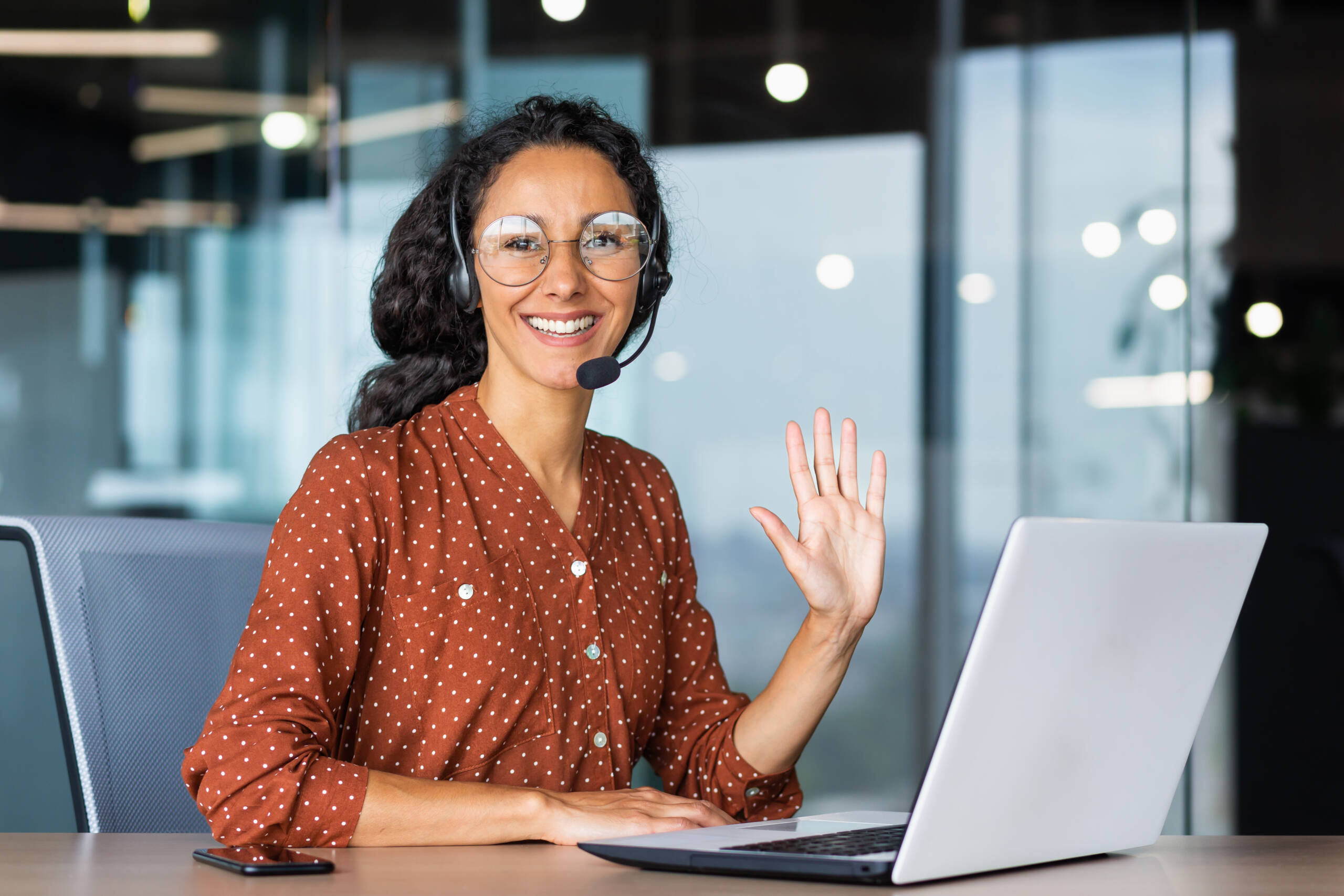 A virtual assistant with a headset smiling and waving while working on a laptop.